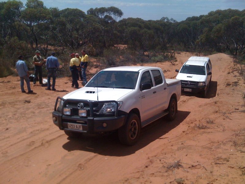 Two white four-wheel-drive utes on a red sand track with a group of workers nearby