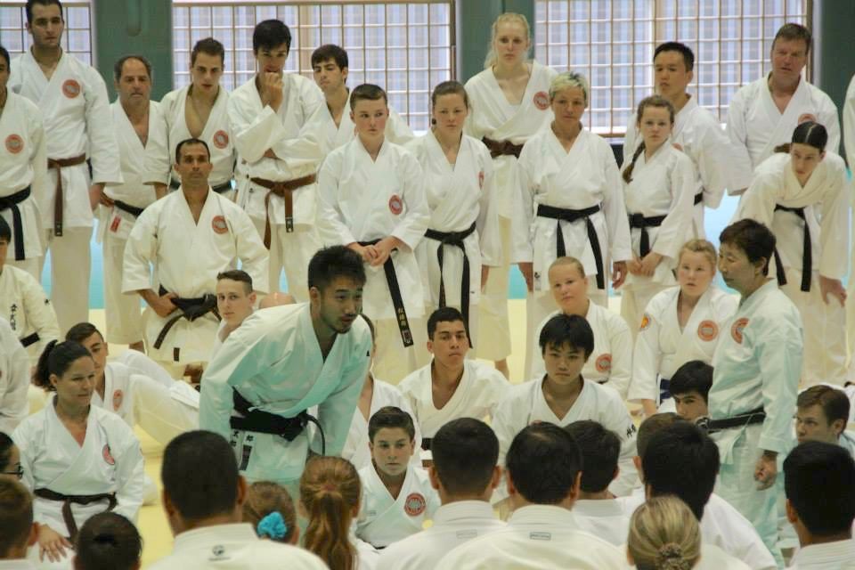 A WSKF instructor leading a large class, with students in white gi and coloured belts watching on the mat
