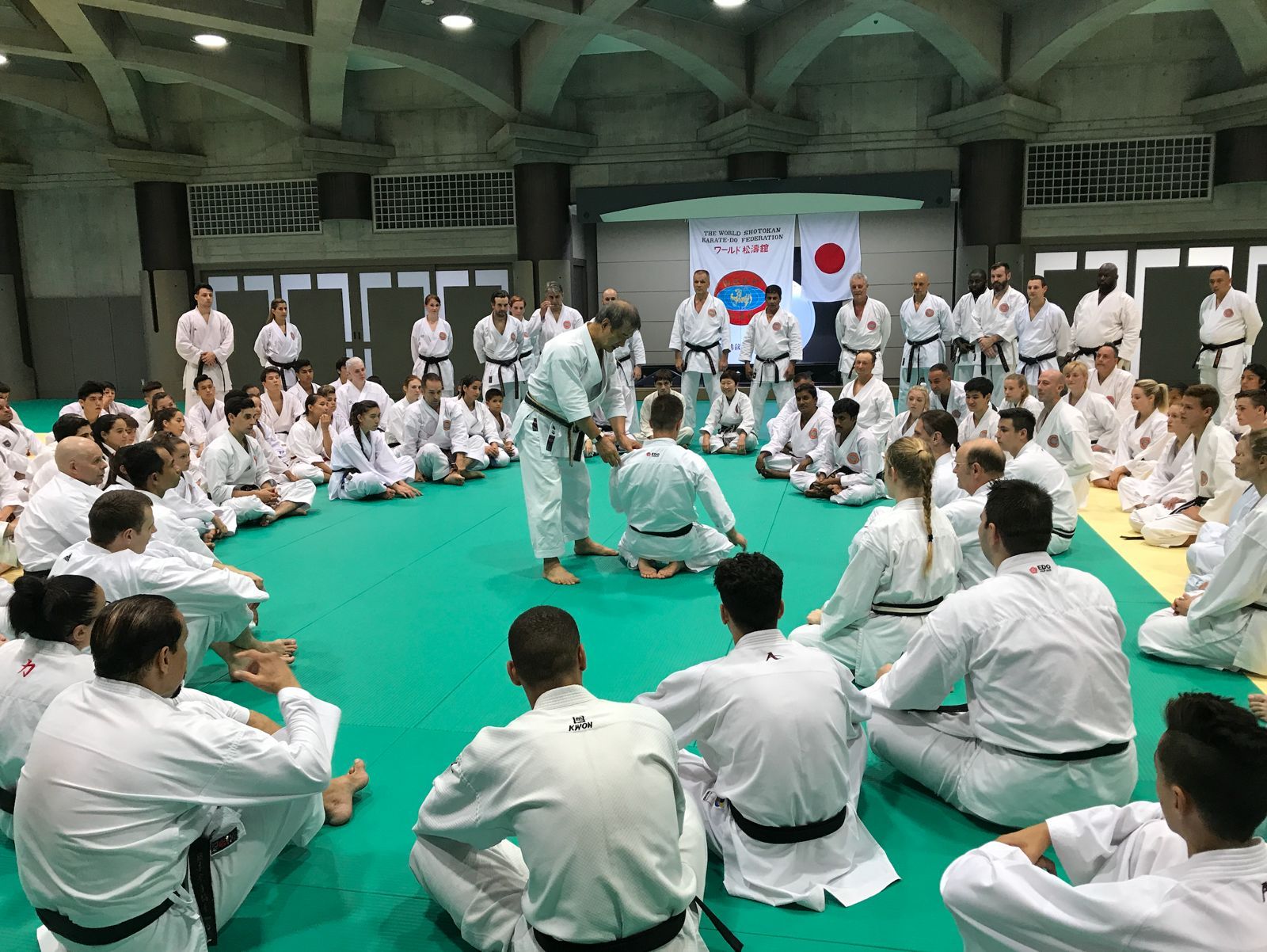 A senior instructor adjusting a kneeling black belt student in front of the World Shotokan Karate-do Federation banner and the Japanese flag, with students seated around the mat